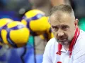 Skupiony Nikola Grbić, trener siatkówki, w białej koszulce z czerwonymi akcentami, na tle żółto-niebieskich piłek.
