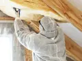 Pracownik w kombinezonie montuje wełnę mineralną na poddaszu. To kluczowy etap prac, wpływający na koszt ocieplenia poddasza i komfort cieplny.