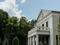 Pałac Grąbkowo, biały budynek z kolumnami i balkonem, otoczony bujną zielenią drzew pod błękitnym niebem z chmurami.