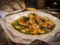 Merluza a la gallega servida en plato blanco con patatas, guisantes y perejil. Pan rústico en cesta al fondo.