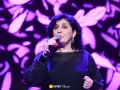 Kobieta śpiewa na scenie, w czarnej, ozdobnej sukni. Wiek męża Eleni nie jest widoczny na zdjęciu.