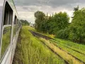 Widok z okna pociągu na tory kolejowe otoczone zielenią i chmury na niebie.