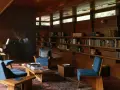 A cozy living room with blue chairs, a piano, and bookshelves. The clerestory windows above let in natural light, illuminating the warm wood interior.