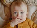 Maluch z niebieskimi oczami wkłada rączkę do buzi, być może przez ząbkowanie. Jak pomóc dziecku w tym trudnym okresie?