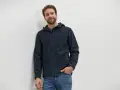 Mann mit lockigem Haar trägt eine dunkelblaue Softshelljacke und Jeans. Perfekt für das nächste Abenteuer oder zum Softshelljacke waschen.