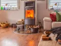 Un poêle bouilleur mixte crépite, réchauffant le salon où un chien dort paisiblement sur un tapis. Bois et paniers complètent ce tableau cosy.