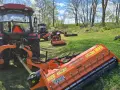 Pomarańczowy mulczer używany, podłączony do traktora, gotowy do pracy na zielonej łące.