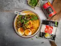 Szybki ramen przepis: miska pełna aromatycznego bulionu z grzybami shiitake, tofu, jajkiem i szczypiorkiem. Obok opakowanie makaronu i grzybów.