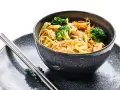 Deliciosa receta con noodles de arroz, brócoli, zanahoria y otros vegetales frescos en un tazón gris, acompañado de palillos.