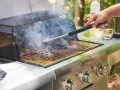 A hand uses a grill brush to clean the grates of a smoking Saber grill, showing the best way to clean a grill for your next cookout.