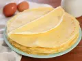 Piatto di crepes senza burro, leggere e dorate, con due uova sullo sfondo.
