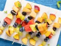 Spiedini di frutta colorati con pesche, melone, anguria, ananas, mirtilli, more e uva, guarniti con menta fresca.
