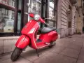 Czerwony skuter Vespa, idealny do zdobycia prawa jazdy AM, stoi na chodniku przed budynkiem.