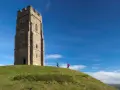 Two hikers ascend the grassy slope towards the iconic Glastonbury Tor tower under a bright blue sky.