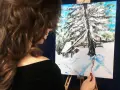 Artystka kończy malowanie zimowego domku w górach. Obraz przedstawia zasypany śniegiem krajobraz z chatką i drzewami.