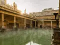 Steaming green water fills the Great Bath, surrounded by ancient Roman ruins with columns and torches.
