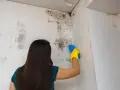 Mujer con guantes amarillos limpiando moho de la pared con un spray.