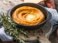 Un cremoso puré de zanahoria en un cuenco negro, coronado con mantequilla derretida. Romero fresco y granos de pimienta añaden aroma.