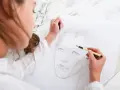 Konstnär i arbete med en ansiktsteckning i mixed media. Skisser av kroppar syns i bakgrunden.
