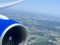 View from an airplane window, showing a blue engine and wing, with a patchwork of green fields and a small town below. Perfect for day trip flights.
