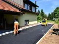 Pracownik równa łopatą świeży asfalt na podjazd, a walec go ubija. Dom w tle.