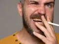 Mężczyzna z zarostem trzyma papierosa, odsłaniając żółte zęby palacza.