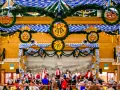 Wnętrze namiotu na Oktoberfest ozdobione niebiesko-białymi girlandami i wiankami z precelkami. Ludzie siedzą przy stołach, a w tle widać orkiestrę.