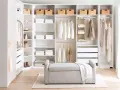 A bright, organized closet with white DIY closet shelves, featuring neatly folded clothes, baskets, and hanging garments. A grey bench sits in front.