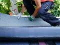 Handwerker verlegt Dachschindeln. Eine Rolle Bitumenbahn liegt bereit, während die neue Schicht auf dem Dach ausgebreitet wird.