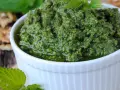 Pesto z pokrzywy w białej miseczce, obok orzechów włoskich i świeżych liści pokrzywy.