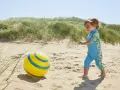 Chłopiec w niebieskim stroju kąpielowym z neoprenu bawi się żółtą piłką na plaży.
