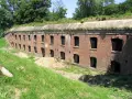 Długi, ceglany mur z licznymi otworami okiennymi i drzwiami, część forty twierdzy przemyśl, porośnięty trawą i zielenią.