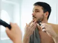 Hombre con barba corta se afeita la barba con una recortadora, buscando un afeitado apurado frente al espejo.