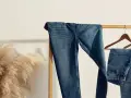 Varios pares de pantalones de **tejido denim** cuelgan de una percha de madera. A la izquierda, un gran ramo de hierba de la pampa añade un toque natural.
