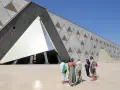 Grupa turystów podziwia nowoczesne muzeum w Gizie, którego fasada zdobiona jest hieroglifami i geometrycznymi wzorami przypominającymi piramidy.