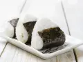 Trzy japońskie kulki ryżowe, onigiri, owinięte w ciemne wodorosty nori, ułożone na białej, ozdobnej tacce.