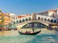 Una gondola solca il Canal Grande a Venezia, passando sotto il Ponte di Rialto, con edifici storici e altre imbarcazioni sullo sfondo.