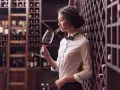 Kto to sommelier? Kobieta w białej koszuli i muszce wącha wino w piwniczce.