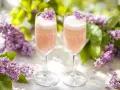 Two champagne flutes filled with pink bubbly drinks, garnished with purple lilac flowers.