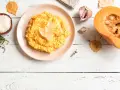 Un cremoso risotto de calabaza, decorado con láminas de queso, junto a ingredientes frescos como arroz, tomillo, ajo y una calabaza partida.