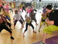 Grupa kobiet ćwiczy podstawowe kroki zumby w sali gimnastycznej, naśladując ruchy instruktora.