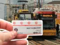 Warszawski bilet 20 minutowy w dłoni, na tle tramwajów i miejskiej zabudowy.