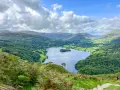 A stunning view of a lake surrounded by green hills and trees, perfect for day trips from Liverpool. A lone hiker walks down a path in the foreground.