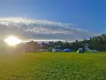 Słońce przebija się przez chmury nad polem namiotowym. Rozstawione namioty i samochody zapowiadają udany camping.