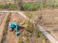 Koparka pracuje przy budowie drogi, która może być częścią służebności drogi. Teren jest częściowo zadrzewiony i porośnięty trawą.