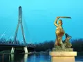 Syrenka warszawska z mieczem i tarczą stoi nad Wisłą, w tle nowoczesny most.