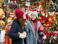 Två kvinnor njuter av julmarknader i Skåne, omgivna av julpynt och ljus. De håller presenter och ler mot varandra.