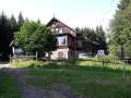 Drewniany budynek schroniska Sowa otoczony lasem. Przed nim zielony trawnik i droga.