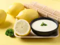 Un bol de sauce beurre citron crémeuse, garni de persil, avec des citrons frais et un rouleau de bambou en arrière-plan.