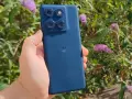 En hand håller en mörkblå motorola edge 60 fusion med fyra kameror mot en bakgrund av gröna blad och lila blommor.